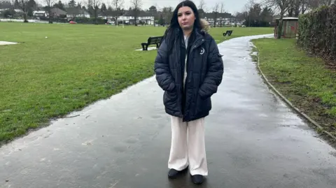 A woman in a park on a grey, overcast day. She is wearing a blue puffa parka jacket with a fluffy hood. 