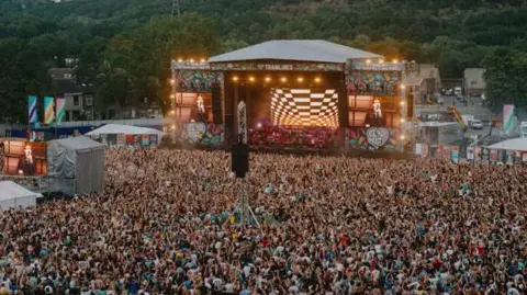 Tramlines/Carolina Faruolo A huge crowd of people at an outdoor concert. A large stage with big screens reveals a tall middle aged man in a brown jacket and dark hair