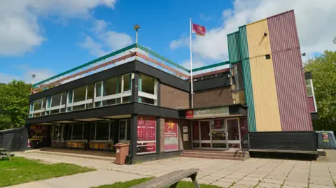 Martin Giles/BBC The outside of Norwich Rugby Club. It is a large brick building with windows and a red flag is raised. There is a blue sky and white clouds above.