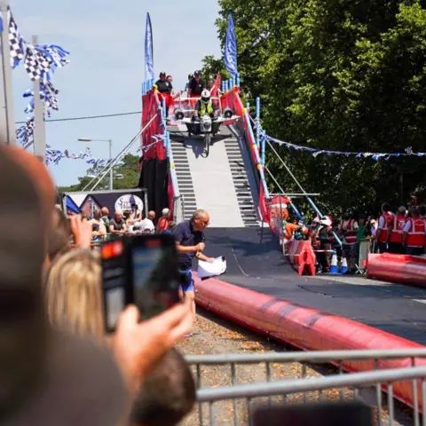 Stuart White A car resembling a plan rolling down the ramp. It has red barriers and blue flags on top. It is beside a large tree, and is nearly half the height of it.