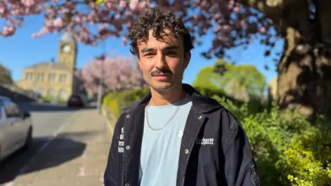 Man with dark curly hair and a mustache wearing a dark blue jacket, light blue top and a silver chain.
