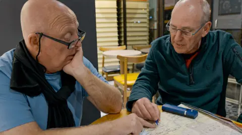 BBC/Studio Lambert Melvyn and Brian discussing their route in a cafe. Both are pointing at a location on a large map on the table.