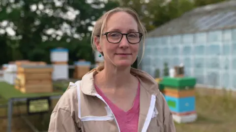 Gwyndaf Hughes/BBC A woman wearing a pink t-shirt and a brown beekeeper suit stands in a field with blue and green bee hives and a greenhouse behind her 