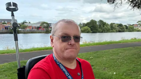 Stephen Magee has short grey hair and is wearing sunglasses, a red T-shirt and a blue lanyard. He is sitting in his mobility scooter in a park, with the River Bann visible in the background.