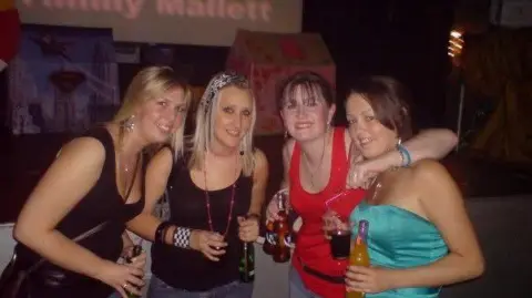 Debbie Travis-Waller, is smiling with three other friends. They are all holding glass bottles of drinks. Mrs Travis-Waller has a straight, dark red hair, pulled back in a ponytail and a fringe. She is wearing a red top and has her left arm round the shoulders of a woman with dark hair and a green strapless top. To her right are the two other women who have longer, blonde hair, both wearing black vest tops. There is a stage behind the group.
