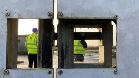 Two men wearing yellow high viz jackets and hard hats. They're standing behind a metal gate.