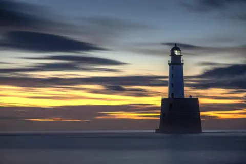 Gina Galbraith A lighthouse in the sea. The sky is orange with dark clouds streaked across it.