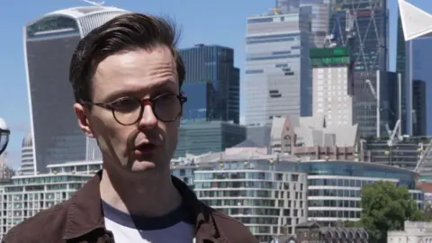 A young man with glasses and brown hair is being interviewed - the skyscrapers of the City of London fill the background of the image. 