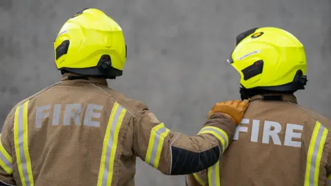 PA Media Two firefighters are facing away from the camera and one has their hand on the other's shoulder. They are wearing brown jackets with hi-vis strips on them and the word fire in capital letters. They are also wearing bright yellow helmets.