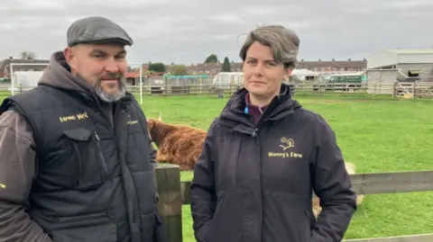 A man and a woman, both dressed in black jackets with yellow "Nunny's Farm" logos, stand in front of a brown Highland cow in a green field.