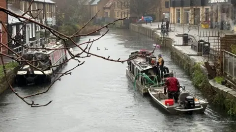 Martin Heath/BBC A motor launch being steered by a man in a red coat and woollen hat and pushing a larger, burnt-out boat which also has a person at the tiller. It is passing a narrowboat to the left, which is moored in front of two-storey brick houses. There are further houses in the distance.