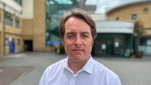 A head and shoulders picture of Thom Lafferty. He is standing in front of a hospital and is wearing a white shirt with the top button undone.