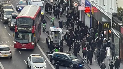 An aerial view of a large group of people on Clapham High Street. A red bus is pulling away while an unmarked police car and a police van are on the road in front of it.