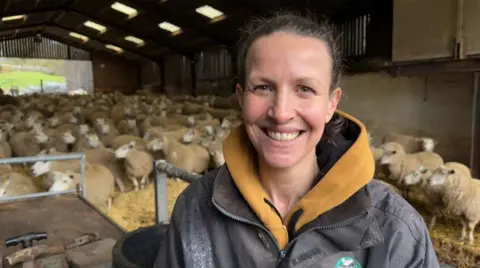 Bryony Gittins, a farmer with brown hair in a ponytail, wearing a black jacket with a yellow jumper. She stands in front of her sheep and is smiling