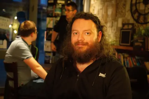 A man with a long beard and dark hair looks at the camera