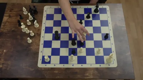 A white and blue chess board with white and black pieces on a table, some pieces on the board and some to the side of it, with hand moving one of them