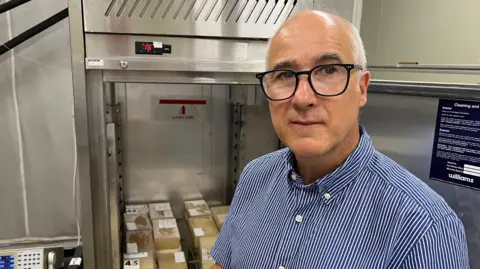 A man in a blue striped shirt stands in front of a stainless steel fridge. The door is open, showing it is full of blocks of cream coloured gel. There are wires coming out of the blocks.