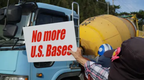 A man holds a white sign with red writing saying "no more US bases". He is wearing a plaid shirt and a cap, but his face cannot be seen. He stands in front of a blue and yellow concrete truck.