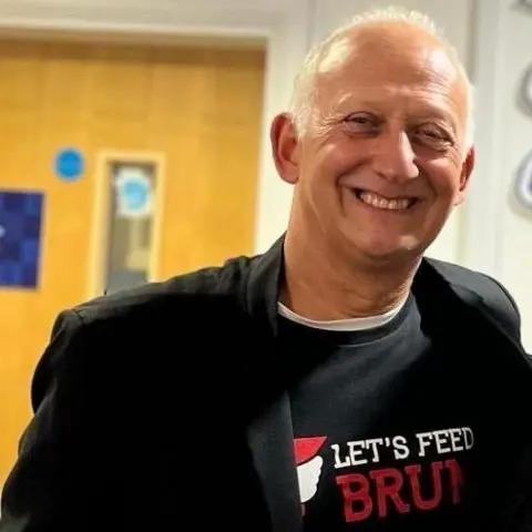Let's Feed Brum A close-up of a man with white hair grinning at the camera. He wears a black t-shirt which says 'Let's Feed Brum.'