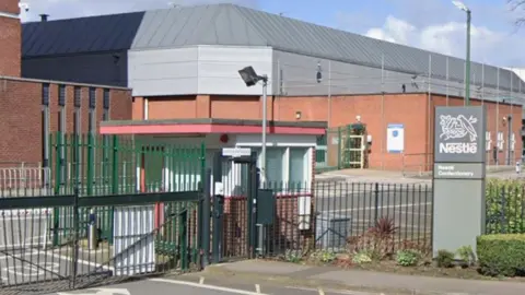 An industrial building behind a green fence and gate, with a gatehouse and a grey column outside with the Nestle logo on it and the words Nestle Confectionery