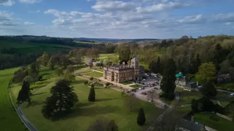 Drone photograph of a mansion, which serves as a private school, surrounded by parkland and woodland in the Cotswolds countryside. There is a car park to the side and manicured lawns surrounding the building.