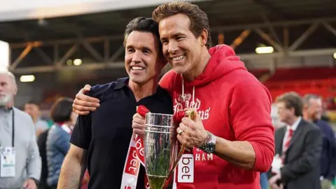 Getty Images Rob, left, and Ryan hold the Championship trophy on the pitch. Rob is wearing a dark-coloured polo shirt and Ryan is wearing a red hoody