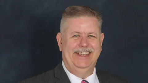 Fife Council John has short grey hair. He is smiling in the picture. He is wearing a grey suit and red tie.