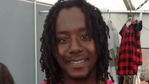 Curtis Green, a  young, black man with chin length hair who is smiling at the camera. Behind him are some clothes hanging off a clothes rail, including a red and black top.