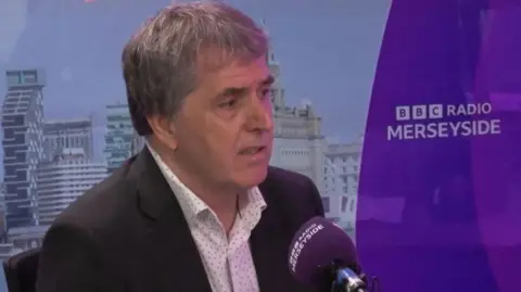 Steve Rotheram is a middle-aged man with dark, greying medium-length straight hair. He is wearing a dark suit jacket with a patterned white shirt that is open at the neck. He is being interviewed in BBC Radio Merseyside's studio - the backgrop and microphone is purple with station branding in white.