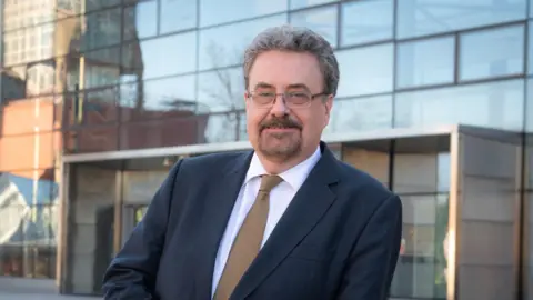 Prof Iain Gillespie in a blue suit and brown tie and wearing glasses, leans on a wall outside a university building
