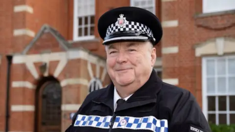 A head and shoulders image of Howard Rayner in his Special police uniform of a black jacket with a strip of blue and white squares across the chest, and a police issue black peaked cap with the Essex Police badge in the centre. He is standing in front of a red brick Victorian building.