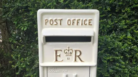 A mounted postbox painted white with the ER II and Post Office lettering painted gold.