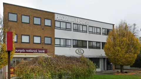 A building that is brown on the left and grey on the right, with the words Hazara House on the outside on the highest storey. Trees are in front and to the side of the building.