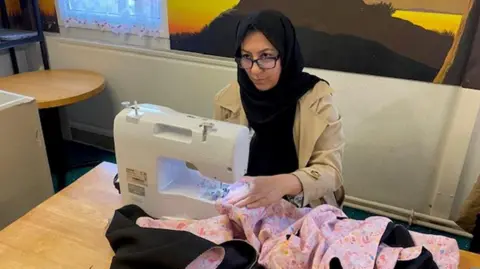 Afghan refugee Aziza wearing glasses sits at a desk uses a sewing machine to make a pink incubator cover.