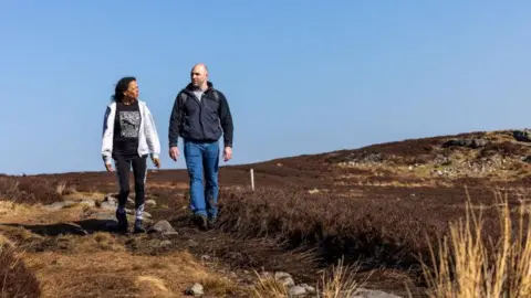 Two hikers traversing a path on the Rothbury Estate. They are a man and a woman and are wearing coats and walking trousers. The ground is made of rough grass and dark coloured plants and the sky is very clear.