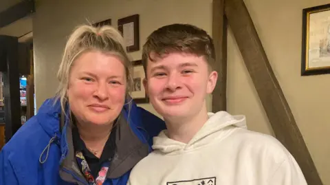 Joshua, on the right, wears a white hoodie sits next to his mum Dawn, on the left. She's wearing a blue coat over a black top with cartoon characters on it.