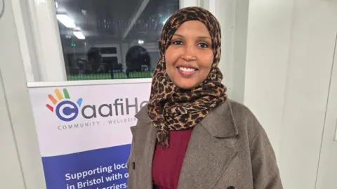 BBC Huda Hajnur smiling at the camera inside a room in a community centre. She is wearing a leopard print hijab, a burgundy top and a brown coat. She is standing in frnt of a CaafiHealth banner.