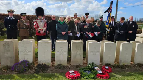 Ministry of Defence A row of white war graves bearing crosses, one of which appears particularly bright white and has poppy wreathes beside it. Several people are lined up behind the row of grave, including some in military uniform.