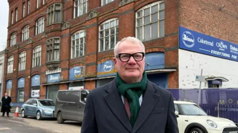 Richard Parker, with white hair and black glasses, is standing near the site. He is wearing a dark top and green scarf and cars are parked outside the building behind him.