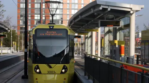 A yellow Metrolink tram at a platform. It has Eccles via MediaCityUK on the electronic indicator board
