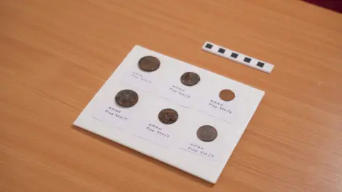 Royal Navy Museums A close up of five 19th century coins and a token on a piece of paper, each labelled. The paper is on a wooden surface.