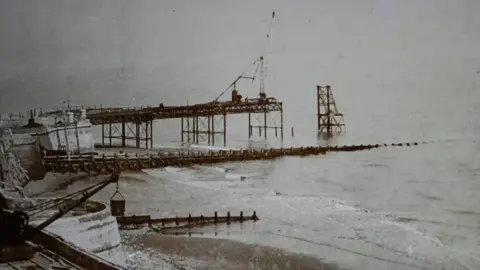 Cromer Museum A black and white image of a pier being constructed. Iron framework stands in the sea, with the beginning of long platform across the top. A crane structure stands at the far end 