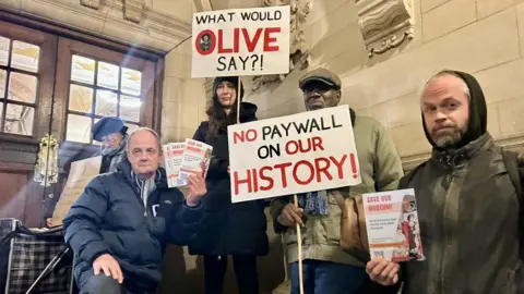 Five protestors from Save Our Museum outside Oxford Town Hall in November. They are holding signs which say "what would Olive say?!" and "No paywall on our history!"