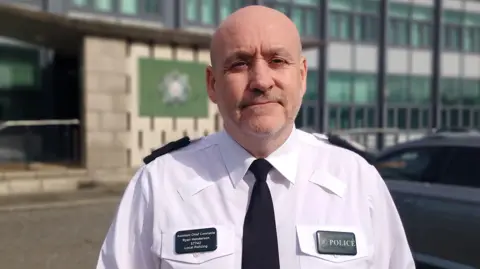 Assistant chief constable Ryan Henderson pictured outside a police station in police uniform, a white shirt and dark green tie with badges saying his name and POLICE. Ryan is bald with grey stubble. 