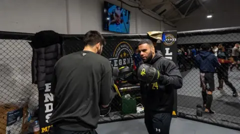 MMA Legends Two men spar inside a miniature octagon cage. Both wear black and one has his back to the camera. The man facing the viewer wears large black sparring gloves and adopts a fighting pose, ready to deliver a strike.