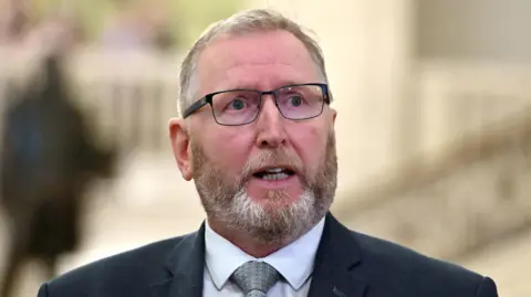 PA Media Doug Beattie. He has short grey-ish hair and beard, wearing glasses, a dark blazer, white shirt and grey tie.