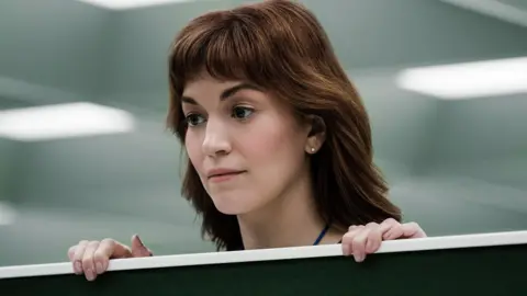 Apple The actor Britt Lower standing in an office. Behind her we can see flourescent lights typical of an office setting, and she is standing with her head poking out above a divider between two seats. She has long red hair and a neutral expression.