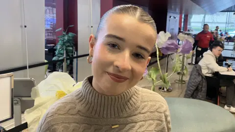A girl with dark hair tied back off her face is looking into the camera. She has a beige jumper on and is wearing large hoop, gold earrings. In the background are people seated at tables.