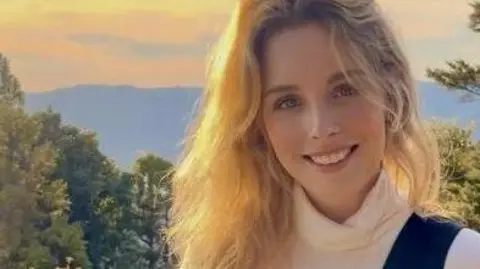 Kim van Daalen Kim van Daalen has long blonde hair and is wearing a white turtleneck jumper and a navy vest. She is in an area which is mainly woodland.