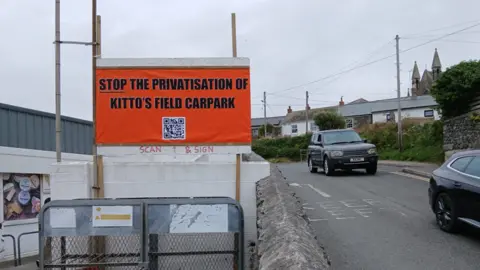 A wide shot of a bright orange road-side sign which reads "Stop the privatisation of Kitto's Field car park".
